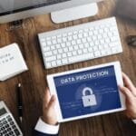 Person holding tablet with data protection lock icon, office desk setup.