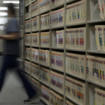 A blurred figure walks past rows of colour-coded medical records files in a hospital records room