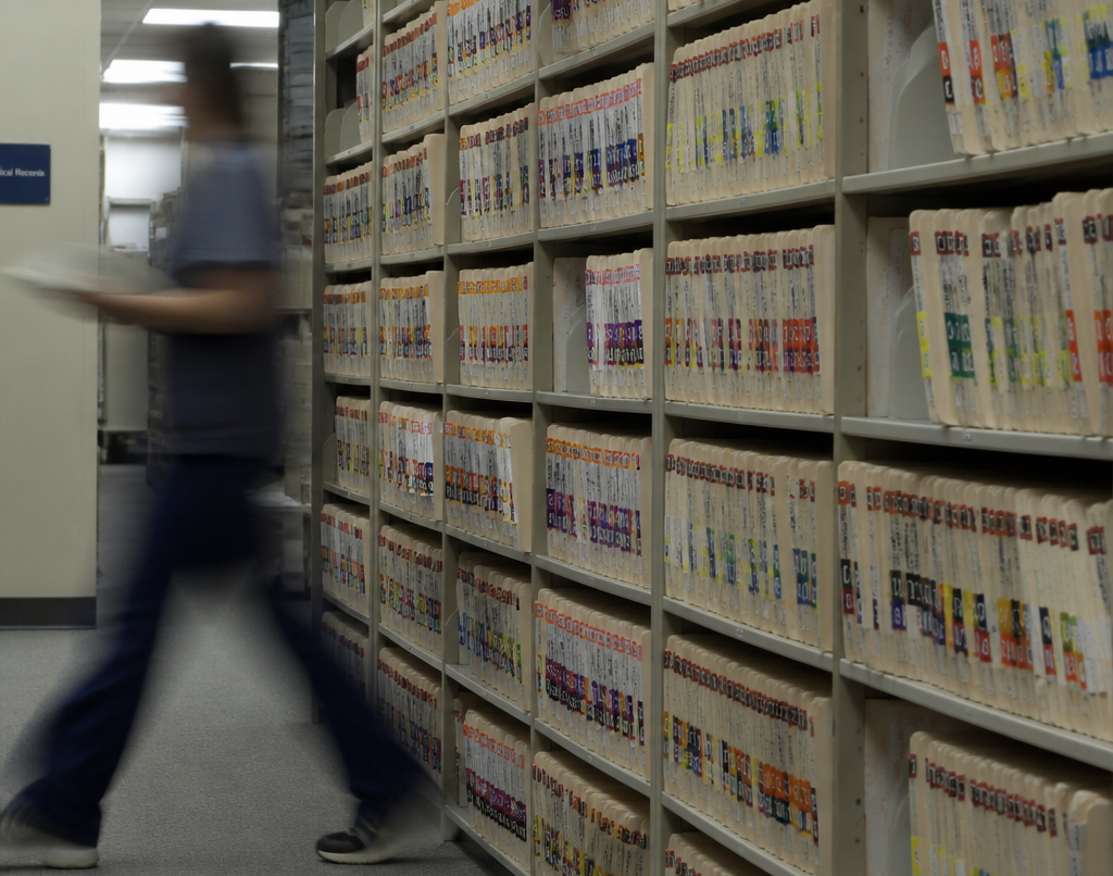 A blurred figure walks past rows of colour-coded medical records files in a hospital records room