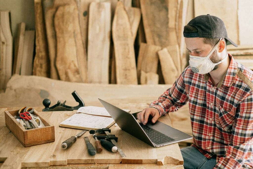 A carpenter wearing a dust mask uses a laptop at a workbench surrounded by woodworking tools.