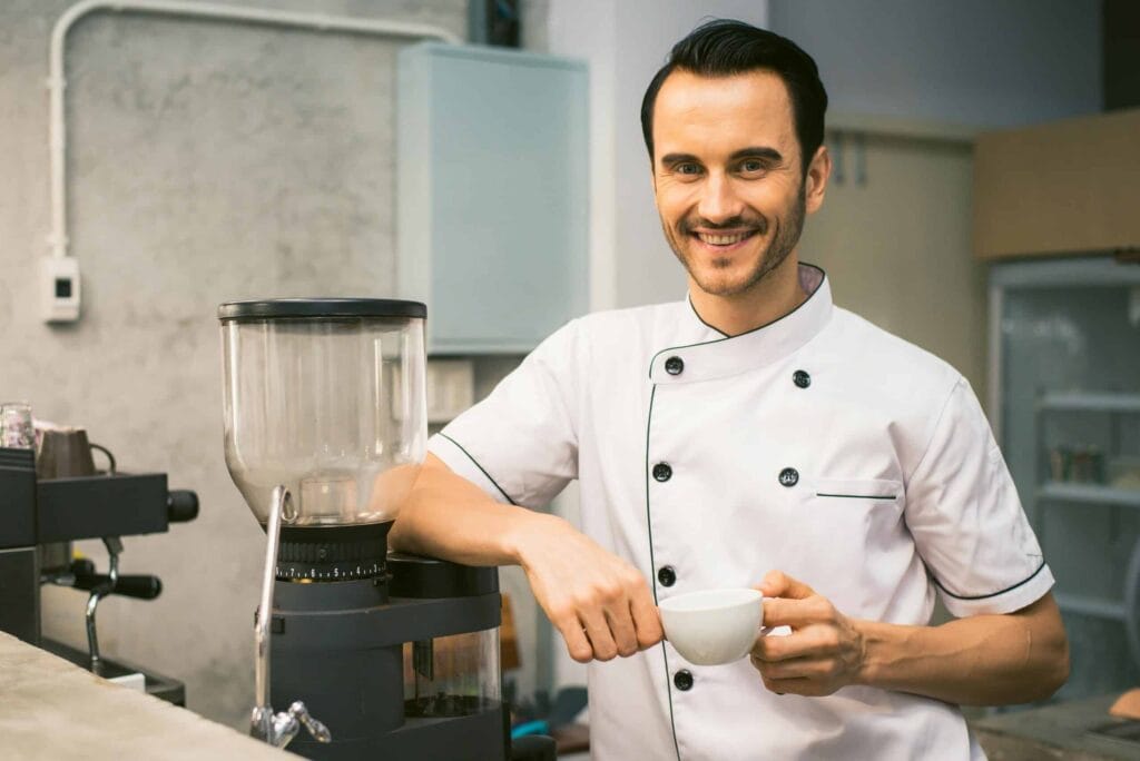 A smiling chef in a white uniform preparing coffee at a bean grinder.
