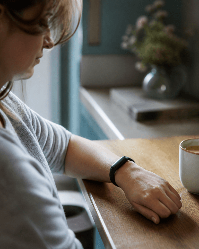 Close-up of a woman's wrist wearing a fitness tracker at a kitchen table, illustrating everyday wearable data collection