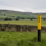 Yellow and black pipeline warning marker post in a grassy field with dry stone walls and Peak District hills behind, marking the route of the proposed Peak Cluster CO2 pipeline