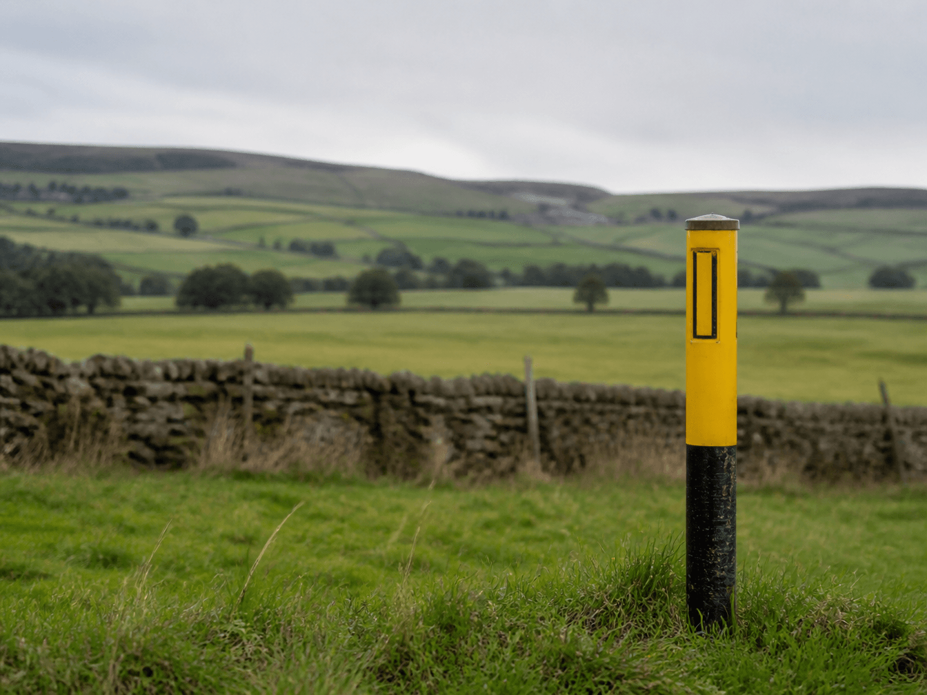 Yellow and black pipeline warning marker post in a grassy field with dry stone walls and Peak District hills behind, marking the route of the proposed Peak Cluster CO2 pipeline
