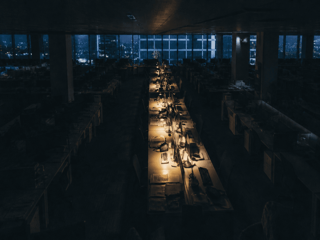 A darkened open-plan office at night, every desk empty and unlit except a single row still working under lamplight, city skyline visible through the windows.