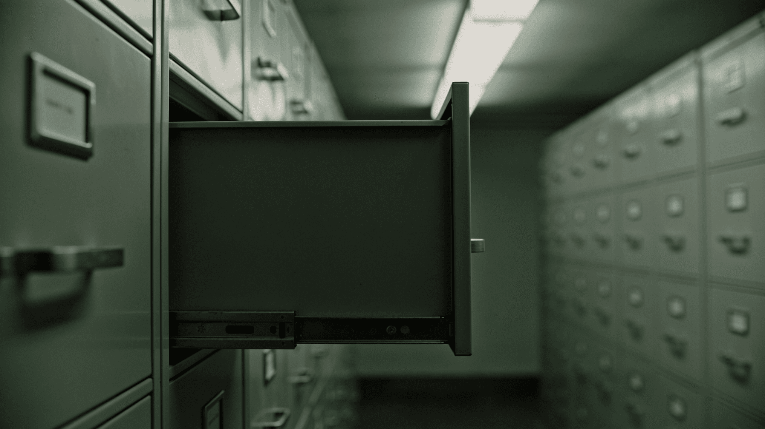 An empty drawer pulled open from a long row of grey metal filing cabinets in a dimly lit storage room