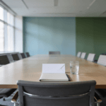 An empty chair at the head of a long boardroom table, with an unopened envelope, a notebook and a glass of water set out in front of it.
