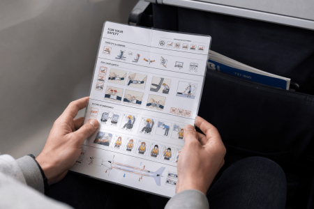 Passenger holding a folded airline safety instruction card, illustrating seatbelt use, brace position, emergency exits, and life vests