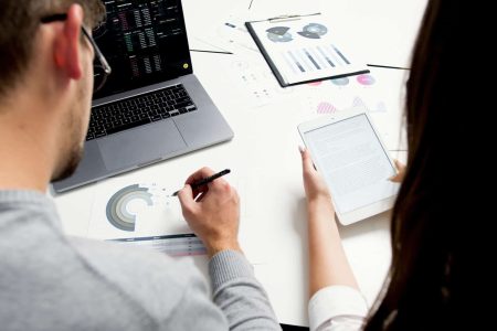 Two people reviewing printed data charts and a tablet, with a laptop showing analytics, during a risk assessment discussion.