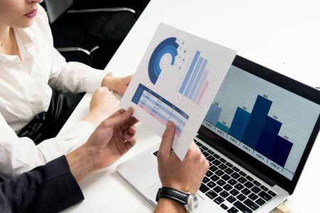 Two people reviewing a printed bar chart and donut chart alongside a laptop showing matching data, during a risk analysis discussion.
